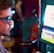 A man wearing sunglasses is intently focused on a slot machine, with colorful lights and graphics illuminating the screen. The scene is set indoors, likely in a casino or gaming area. His attire includes a patterned shirt and a blazer.