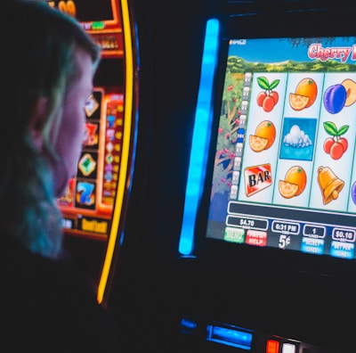 A person is closely observing a digital slot machine with colorful fruit symbols, bars, and a bell. The slot machine screen displays symbols like oranges, plums, cherries, and clouds. The background is dimly lit, creating a focused and somewhat intense atmosphere.