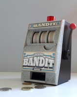 A small, vintage-style slot machine with the word 'Bandit' prominently displayed. It features a lever on the side with a red knob and three reels with a generic design. Several coins are scattered on the white surface in front of it.