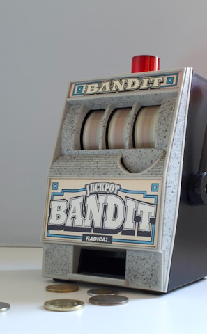 A small, vintage-style slot machine with the word 'Bandit' prominently displayed. It features a lever on the side with a red knob and three reels with a generic design. Several coins are scattered on the white surface in front of it.